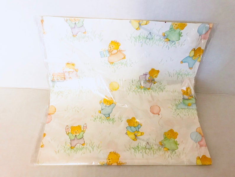 May include: White wrapping paper with a repeating pattern of teddy bears wearing different colored clothing and holding balloons. The bears are playing on a seesaw and holding presents.