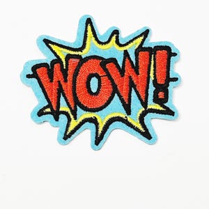 May include: Embroidered patch in a comic book style. The patch features the word "WOW!" in red, outlined in black, set against a yellow and black burst on a light blue background. The patch is likely for clothing or accessories.