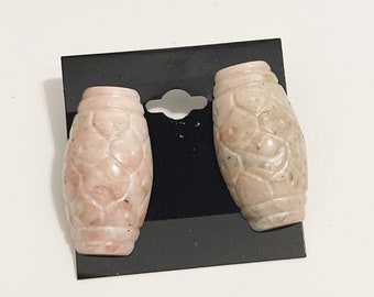 May include: A pair of pink stone earrings with a carved design. The earrings are oval shaped and have a smooth, polished finish.