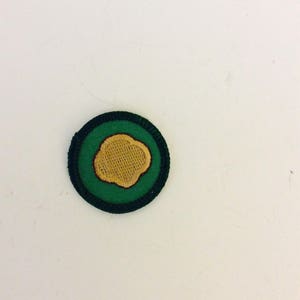 May include: A green and gold embroidered patch with a flower design. The patch has a black border.