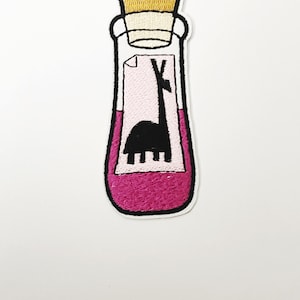 Può includere: Una toppa ricamata da stirare a forma di bottiglia di vetro rosa con un tappo di sughero. La bottiglia contiene un liquido rosa e una silhouette nera di un lama all'interno.