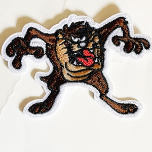 May include: A brown and white embroidered patch of the cartoon character Taz from Looney Tunes. Taz is shown with his mouth open and teeth bared, with a red tongue sticking out. He is in a running pose with his arms and legs outstretched.