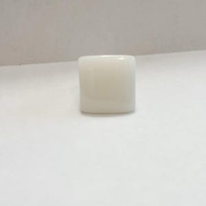 Puede incluir: Un anillo cuadrado de color blanquecino. El anillo es de color sólido y tiene bordes redondeados. El fondo es una superficie blanca lisa.