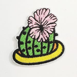 Puede incluir: Parche bordado que representa un cactus verde con puntos negros, una flor rosa y una base amarilla. El parche tiene un borde blanco y está diseñado para su aplicación en ropa o accesorios.