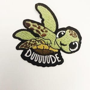 Puede incluir: Parche bordado con una tortuga marina de dibujos animados. La tortuga tiene un cuerpo verde con manchas marrones, un vientre amarillo y ojos grandes. La palabra "DUUUUDE" está bordada en blanco sobre un borde negro.