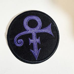 May include: A circular embroidered patch featuring a black background with a purple Prince love symbol stitched in the centre. The design is enclosed within a thick black stitched border, creating a clean and defined edge for the circular shape.