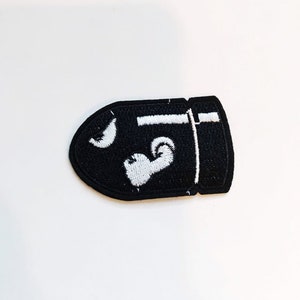 Puede incluir: Parche bordado negro con forma de bala con detalles blancos. El parche presenta una luna creciente blanca y un coraz&oacute;n blanco.