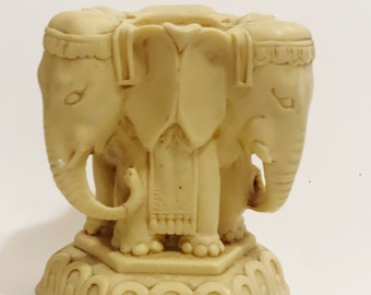 May include: A white ceramic figurine of three elephants standing on a base with a decorative design. The elephants are facing forward with their trunks raised.