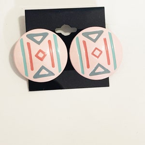 Puede incluir: Un par de pendientes redondos de color rosa con un diseño geométrico en tonos grises, azul verdoso y coral.