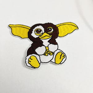 May include: An embroidered iron-on patch featuring a brown and yellow cartoon creature with large ears and big eyes. The creature is sitting with its legs crossed and has a white belly.