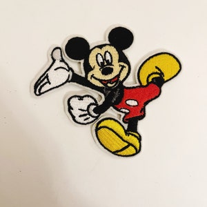 May include: A cartoon embroidered patch of Mickey Mouse in a red and yellow outfit, waving with a big smile.