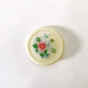 May include: A white button with a rope-like border and a floral design in pink, blue, and green.