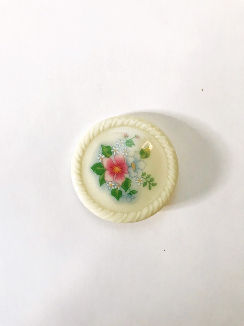 May include: A white button with a rope-like border and a floral design in pink, blue, and green.