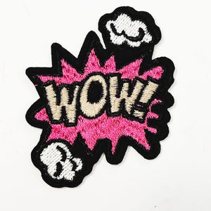 May include: An embroidered patch featuring the word "WOW!" in beige, set against a pink burst design with a black outline. Includes white cloud and skull accents. Ideal for clothing.