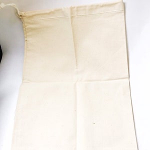 Może przedstawiać: Otwarta, kremowa torba ze sznurkiem wykonana z pł&oacute;tna. Torba jest prostokątna z zszytą g&oacute;rną krawędzią i zamknięciem na sznurek. Tkanina wydaje się być naturalną, niebieloną bawełną lub podobnym materiałem. Torba jest pusta i leży płasko.