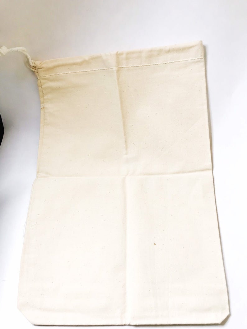 Może przedstawiać: Otwarta, kremowa torba ze sznurkiem wykonana z pł&oacute;tna. Torba jest prostokątna z zszytą g&oacute;rną krawędzią i zamknięciem na sznurek. Tkanina wydaje się być naturalną, niebieloną bawełną lub podobnym materiałem. Torba jest pusta i leży płasko.