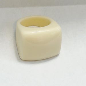 May include: A square ring in a creamy off-white hue. The ring features a smooth, polished finish and a rounded interior. The design is solid and chunky, with a minimalist aesthetic.