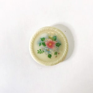 May include: A white ceramic button with a floral design. The button features a pink flower with blue and white flowers surrounding it. The button has a rope-like border.