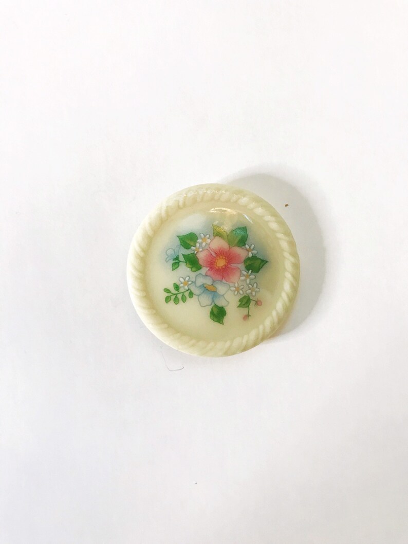 May include: A white ceramic button with a floral design. The button features a pink flower with blue and white flowers surrounding it. The button has a rope-like border.