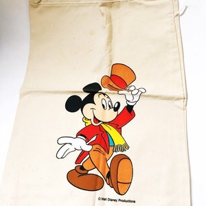 Może przedstawiać: Beżowa torba ze sznurkiem z kolorową ilustracją Myszki Miki w czerwonej kurtce, brązowych spodniach i ż&oacute;łtym szaliku. Miki trzyma pomarańczowy kapelusz. Torba jest zadrukowana napisem "Walt Disney Productions" i "Made in USA".