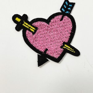May include: An embroidered patch featuring a pink heart pierced by a yellow arrow with a black tip and a blue fletching. The heart is outlined in thick black thread, creating a bold contrast against the pink center and the yellow arrow details.