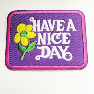May include: Purple embroidered patch with a pink border. The patch features a yellow flower with a green stem and white text that reads "HAVE A NICE DAY".