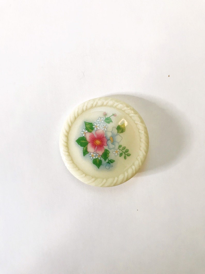 May include: A white ceramic button with a floral design. The button features a pink flower with blue and white flowers surrounding it. The button has a rope-like border.