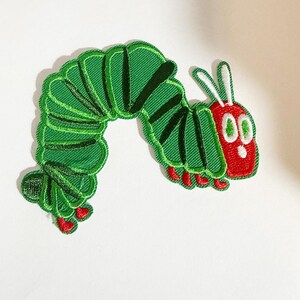 May include: An embroidered patch shaped like a caterpillar with a bright red head, white eyes, and two white antennae. The body is segmented in shades of green with dark green stripes and small red feet along the bottom edge of the curved form.
