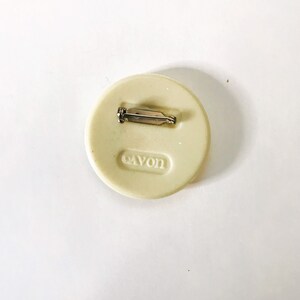 May include: White round pin with a silver pin back. The pin has the word "AVON" embossed on the back.