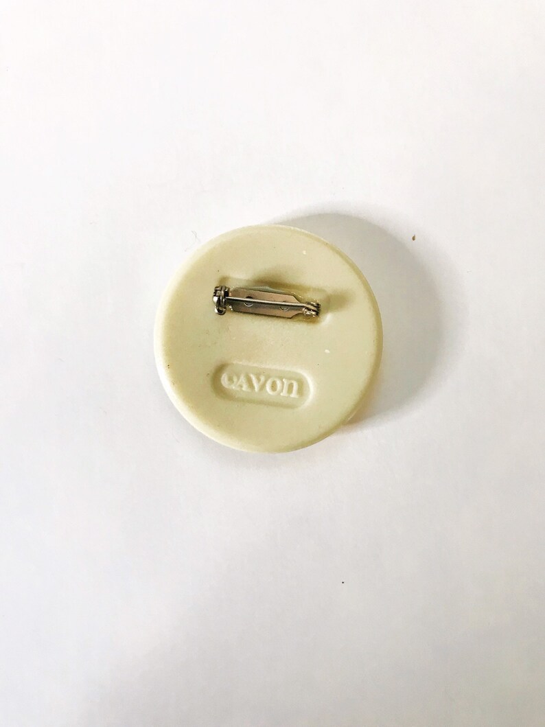 May include: White round pin with a silver pin back. The pin has the word "AVON" embossed on the back.