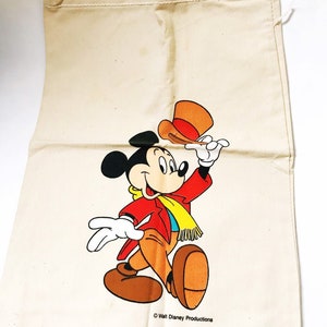 Może przedstawiać: Beżowa torba ze sznurkiem ze kolorową grafiką Myszki Miki w czerwonej kurtce, brązowych spodniach i ż&oacute;łtym szaliku. Miki trzyma pomarańczowy kapelusz. Torba ma napis "Walt Disney Productions".