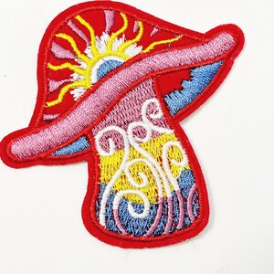May include: Embroidered patch in the shape of a mushroom with a red border. The mushroom cap features a sun design with yellow rays, pink and white accents. The stem has a colorful design with blue, yellow, and pink swirls.