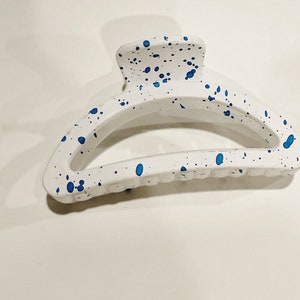 May include: White hair claw clip with blue paint splatter design. The claw clip is a curved shape with a matte finish. The blue paint splatters are randomly placed across the surface of the clip.