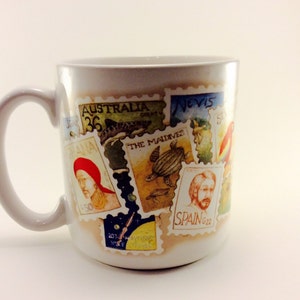 May include: White ceramic mug with a colorful design of postage stamps featuring images of animals, people, and places. The stamps have text that includes "Australia", "The Maldives", "Nevis", and "Spain".