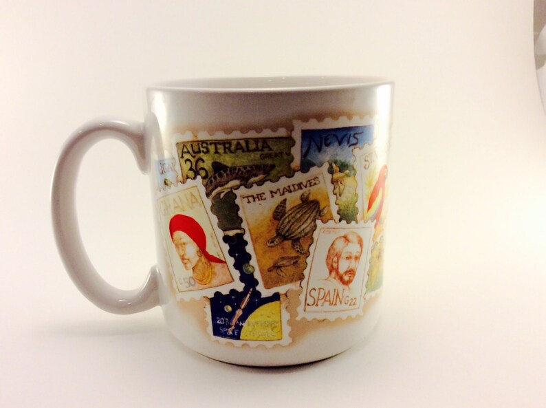 May include: White ceramic mug with a colorful design of postage stamps featuring images of animals, people, and places. The stamps have text that includes "Australia", "The Maldives", "Nevis", and "Spain".
