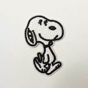 Puede incluir: Parche bordado con Snoopy, el icónico beagle de dibujos animados. El parche es blanco con detalles negros, mostrando la cara sonriente de Snoopy y su pose sentada. Perfecto para ropa o accesorios.
