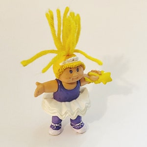 Könnte beinhalten: Eine Cabbage Patch Kid Puppe mit gelben Haaren, die ein lilafarbenes Trikot und einen weißen Tutu trägt und einen gelben Sternstab hält.