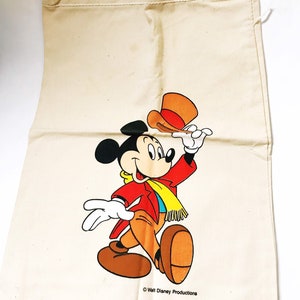 Może przedstawiać: Pł&oacute;cienna torba ze sznurkiem w kolorze kremowym z ilustracją Myszki Miki. Myszka Miki ma na sobie czerwoną kurtkę, brązowe spodnie i ż&oacute;łty szalik, podnosząc pomarańczowy kapelusz. Torba ma napis "Walt Disney Productions" i "Made in USA".