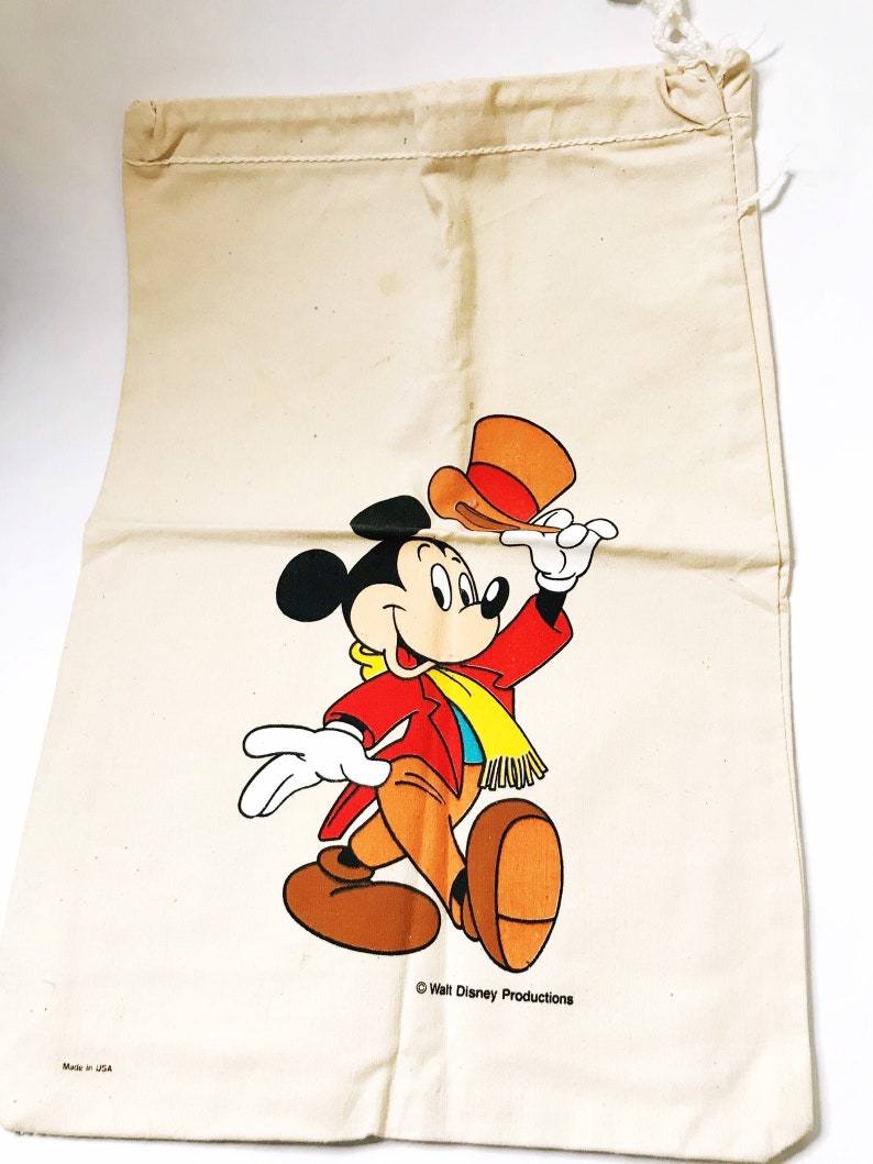 Może przedstawiać: Pł&oacute;cienna torba ze sznurkiem w kolorze kremowym z ilustracją Myszki Miki. Myszka Miki ma na sobie czerwoną kurtkę, brązowe spodnie i ż&oacute;łty szalik, podnosząc pomarańczowy kapelusz. Torba ma napis "Walt Disney Productions" i "Made in USA".