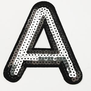 May include: A large, sequined letter "A" patch. The letter is outlined in black felt and filled with silver and gray sequins. The sequins are arranged in rows, creating a shimmering effect. This patch is suitable for clothing or accessories.