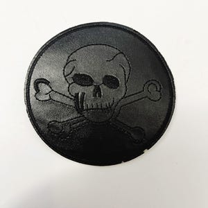 May include: A round, black patch featuring a skull and crossbones design. The skull has detailed eye sockets and a prominent nose. The patch is made of a textured material, likely leather or a similar fabric.