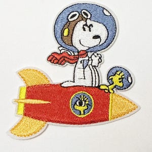 May include: Embroidered patch featuring Snoopy in an astronaut helmet and red scarf, piloting a red rocket with yellow accents. A yellow bird is also on the rocket. The patch has a white border and is on a white background.