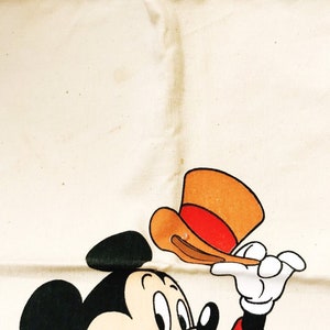 Może przedstawiać: Kremowa torba typu tote z ilustracją kresk&oacute;wkową Myszki Miki. Miki, z czarnymi uszami i czerwoną kurtką, trzyma brązowo-czerwony kapelusz. Torba ma prosty design z przeszytym szwem na g&oacute;rze.