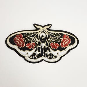 May include: Embroidered moth patch with a black body, white trim, and red rose accents. The patch features intricate detailing and a symmetrical design, suitable for clothing or accessories.