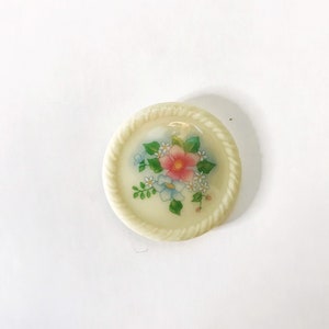 May include: A white ceramic button with a floral design. The button features a pink flower with blue and white flowers surrounding it. The button has a rope-like border.
