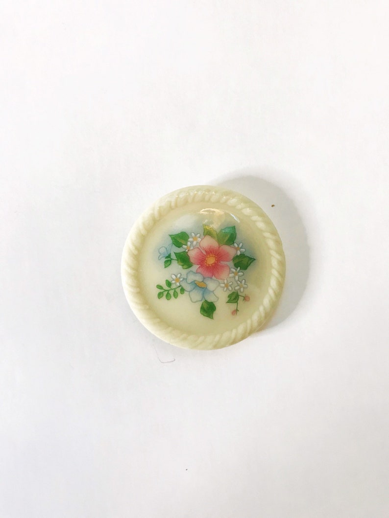 May include: A white ceramic button with a floral design. The button features a pink flower with blue and white flowers surrounding it. The button has a rope-like border.