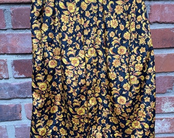 Op de afbeelding: Zwarte rok met een gouden bloemenpatroon. De rok hangt aan een zwarte hanger tegen een bakstenen muur. Het bloemmotief heeft gele en gouden bloemen en bladeren.