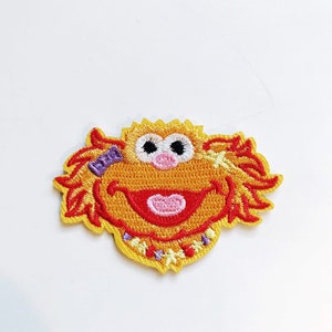 Pode incluir: Um remendo bordado de ferro amarelo e vermelho com um personagem de desenho animado com pêlo laranja, nariz rosa e um grande sorriso. O personagem está usando uma faixa de cabeça roxa e um colar amarelo com flores brancas.