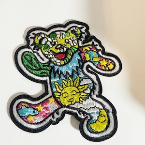 May include: An embroidered patch in the shape of a dancing bear. The bear features a yellow head with white flowers, a blue torso with a yellow sun, and limbs decorated with colourful patterns including stars, clouds, and a crescent moon.