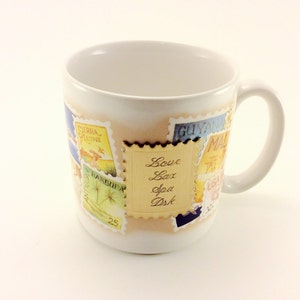 May include: White ceramic mug with a handle. The mug is decorated with colorful postage stamps featuring images of palm trees, birds, and the text "Love Lax Spa Dsk".